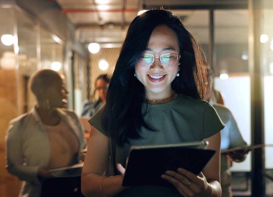 A smiling woman in a modern office setting, holding a tablet and surrounded by colleagues engaged in discussion, symbolizing a social and collaborative work environment.