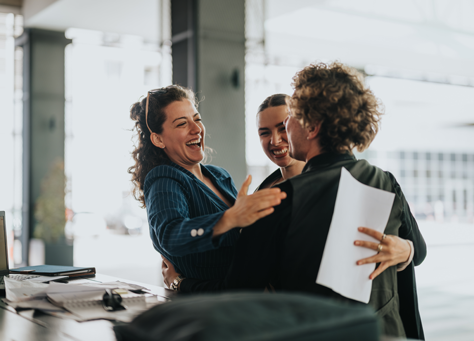 Three colleagues share a joyful moment, hugging and smiling in a professional setting, reflecting the company's strong social culture and emphasis on collaboration, trust, and positive team dynamics.