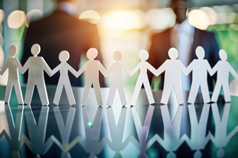 A row of paper cutout figures holding hands, illustrating teamwork and connection, with a blurred business setting in the background, emphasizing the company's commitment to social responsibility.