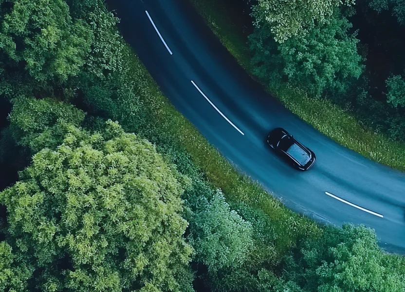 Aerial view of a winding road through a dense green forest with two cars passing. Highlights a sense of harmony between mobility and nature, reflecting a vision of sustainability in modern transportation.