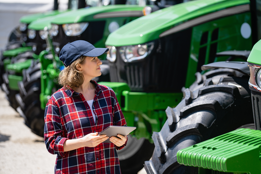 Woman in a checkered shirt and cap holding a tablet while standing in front of a row of green tractors.