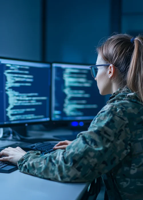 "Woman in military uniform working on cybersecurity tasks at a dual-monitor computer setup, with lines of code visible on both screens."