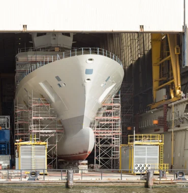 Large vessel under construction inside a covered shipyard hall surrounded by scaffolding and industrial equipment.