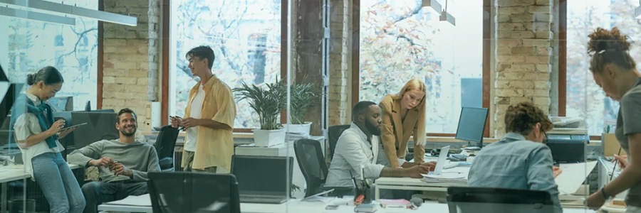 People working and talking in a modern open-plan office with brick walls and large windows.