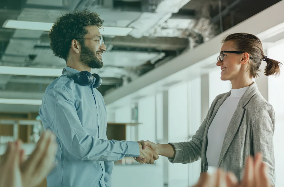 Two colleagues shaking hands in an office environment, with people clapping in the foreground.