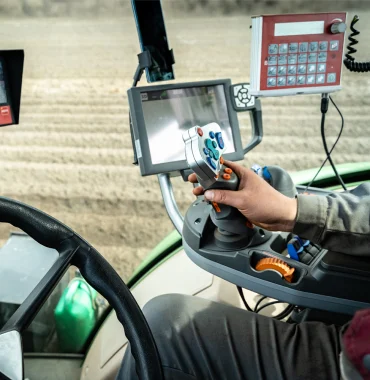 View inside the cabin of agricultural machinery with control panels, steering wheel, and a person operating a joystick on a cultivated field.